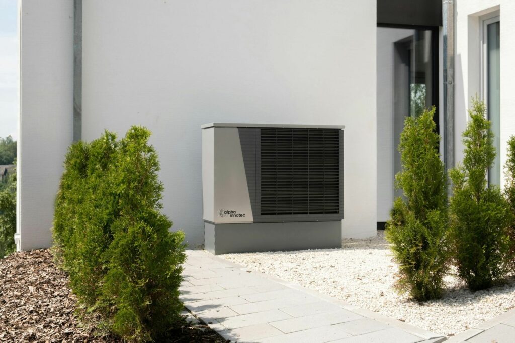 An alpha innotec air conditioning unit installed outside a modern house with green shrubs and a gravel path.