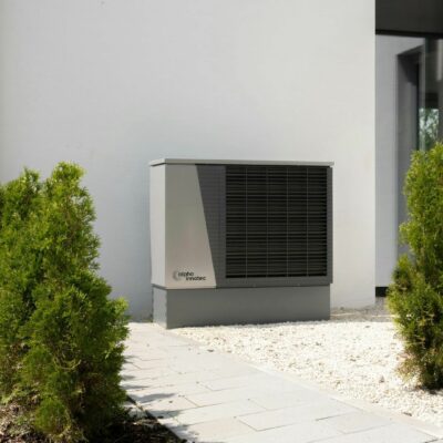An alpha innotec air conditioning unit installed outside a modern house with green shrubs and a gravel path.
